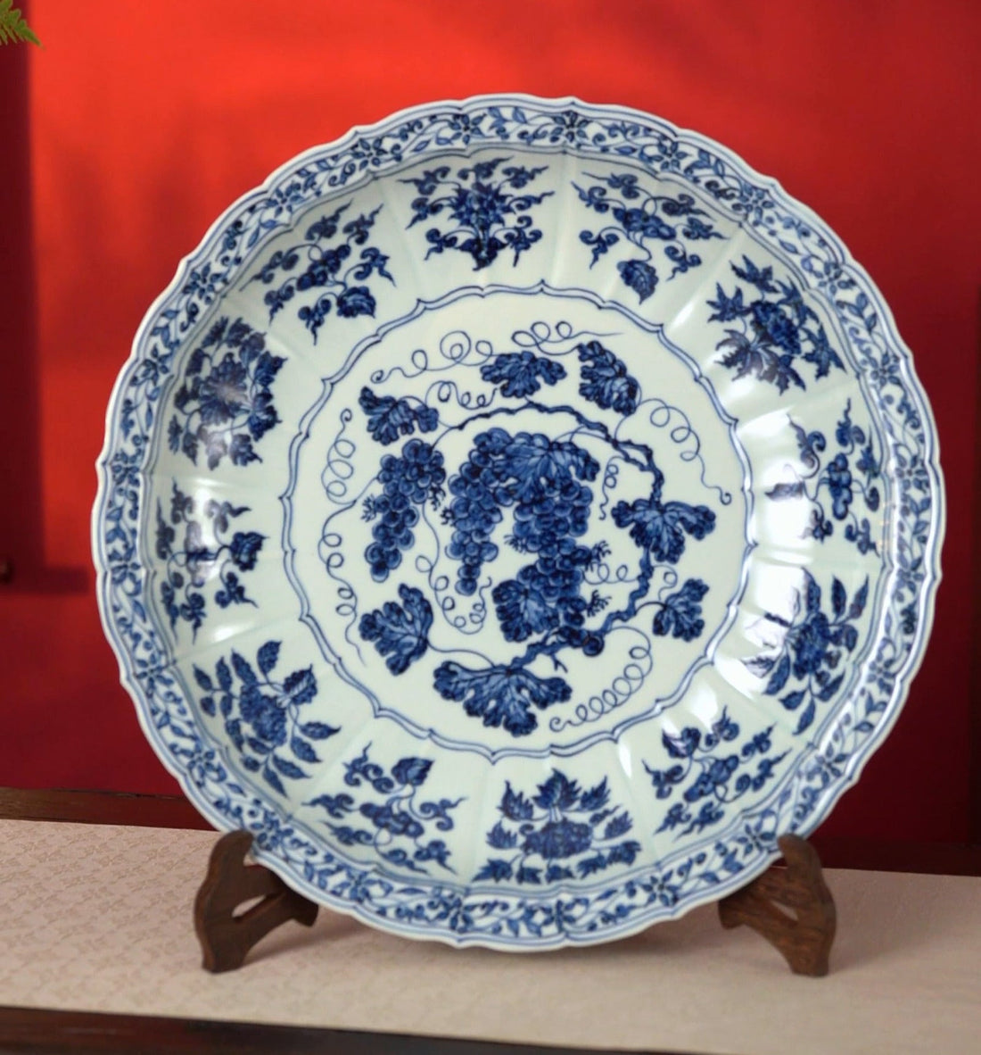 Blue-and-white grapevine plate with a scalloped border, displayed on a wooden stand against a red wall.