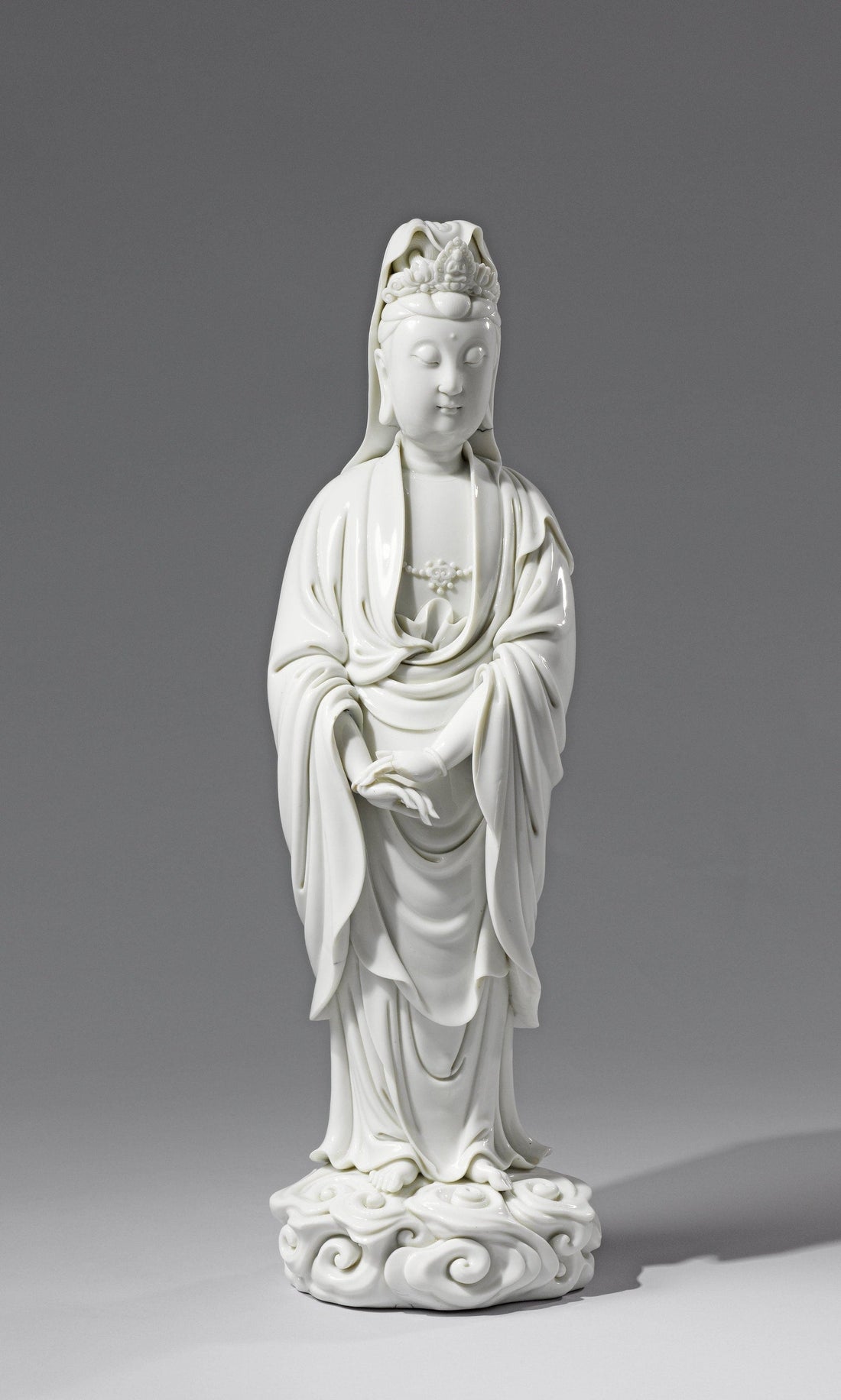 White porcelain Guanyin with lowered eyes, overlapping hands and long robes above a curled base