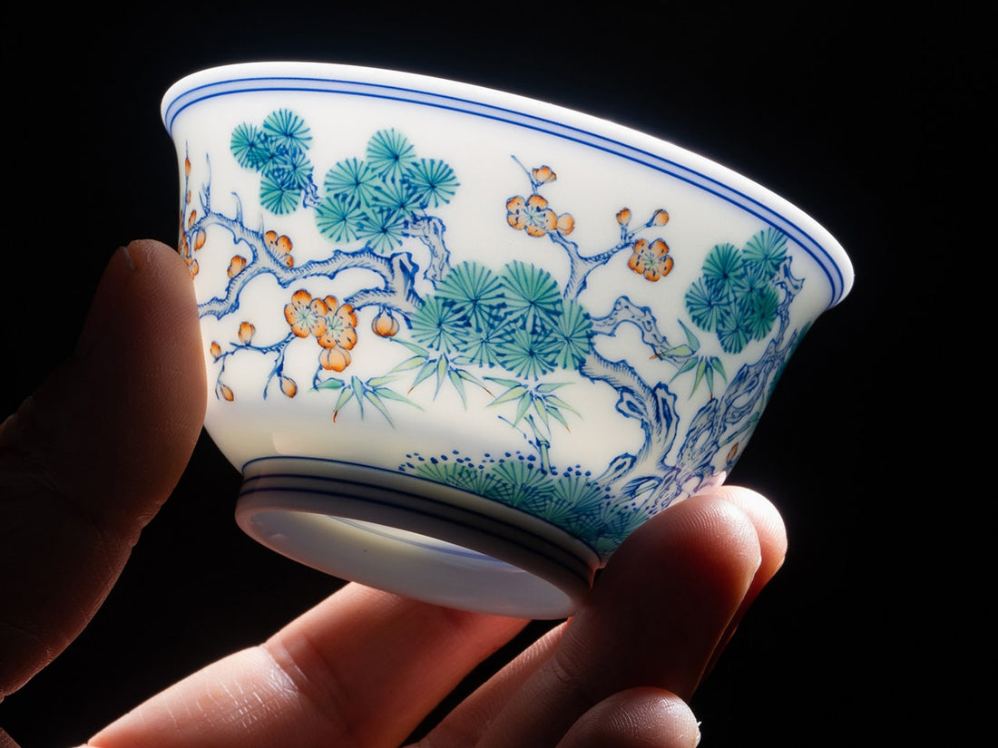 A hand holding a contemporary porcelain teacup with green pine needles, slender bamboo leaves and orange plum blossoms