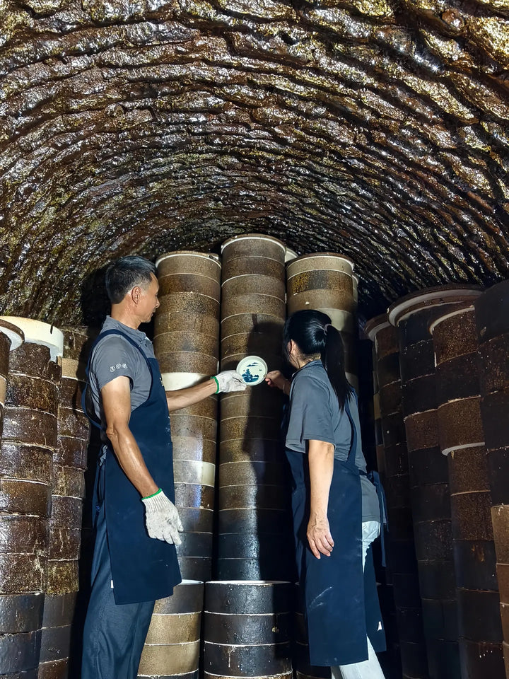 Two people examining a porcelain dish among stacked saggars inside an arched kiln