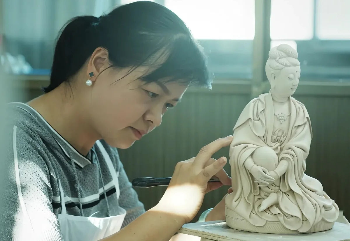 A craftsperson using a hand tool to refine a seated porcelain figure in a sunlit workshop
