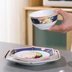 Hand holding the floral rice bowl above two stacked faceted porcelain plates with gold-colored trim.