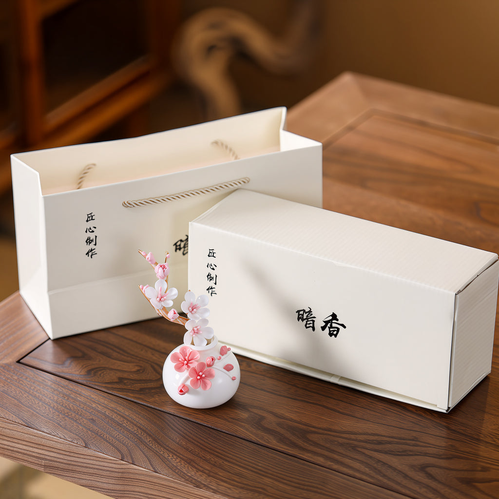 White plum-blossom vase and ceramic branch with a cream-colored gift box and handled paper bag.