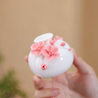 Hand holding a round white porcelain vase with raised pink plum blossoms and buds.
