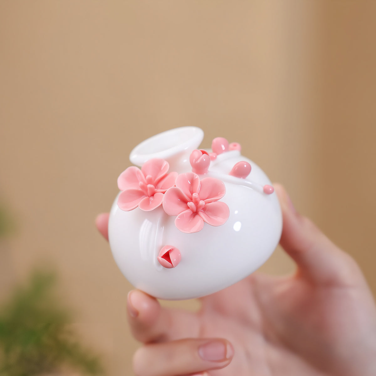 Hand holding a round white porcelain vase with raised pink plum blossoms and buds.