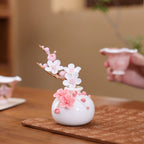 White porcelain plum-blossom vase and ceramic branch on a patterned tea cloth beside teaware.