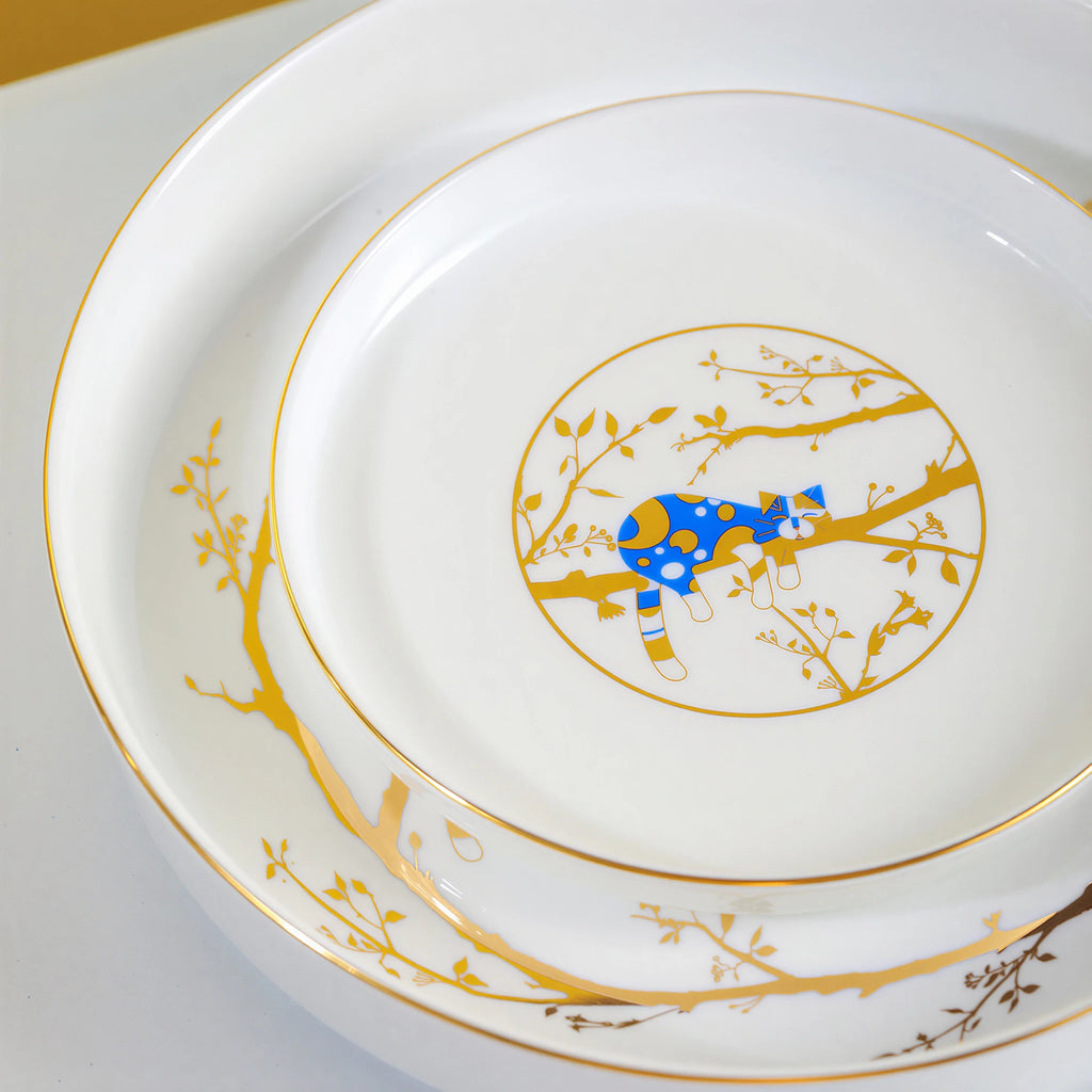 Close view of nested white porcelain plates with a resting blue cat and reflective gold branch decoration.