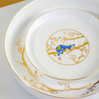 Close view of nested white porcelain plates with a resting blue cat and reflective gold branch decoration.