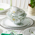 Two botanical porcelain bowls stacked on a plate, showing green stems, pale yellow flowers, and narrow silver-colored rims.
