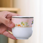 Hand holding the handleless porcelain cup with a blush floral band and narrow gold-colored rim.