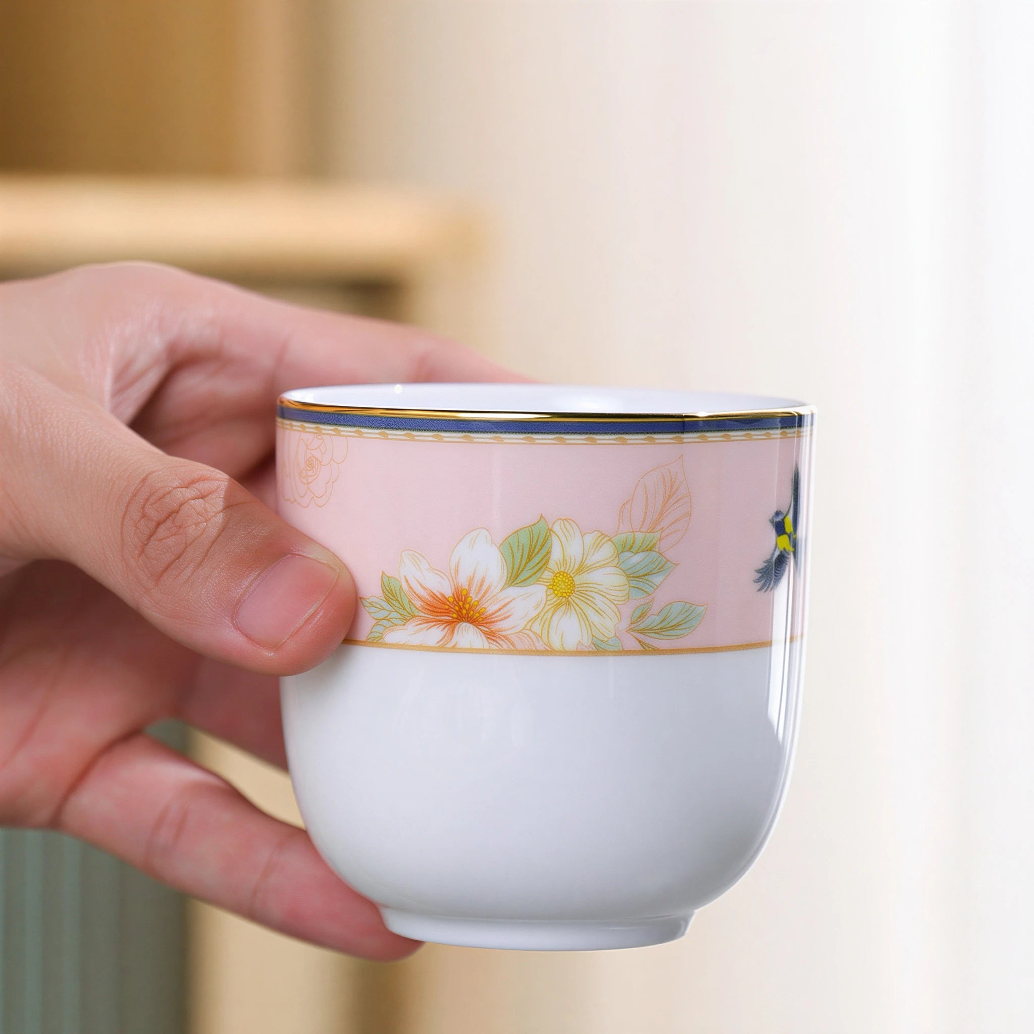 Hand holding the handleless porcelain cup with a blush floral band and narrow gold-colored rim.