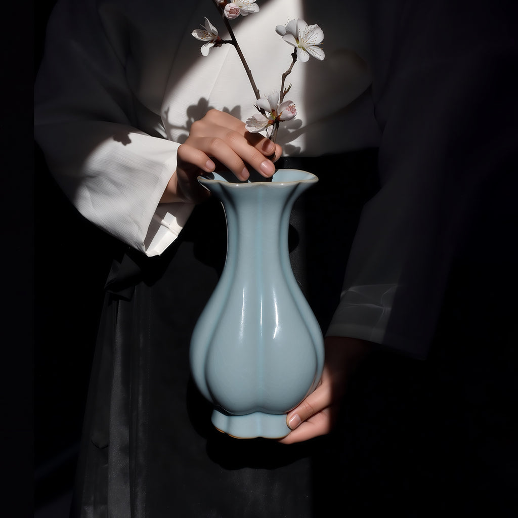 Hands arranging a white flowering branch in a sky-blue Haitang vase against dark clothing.